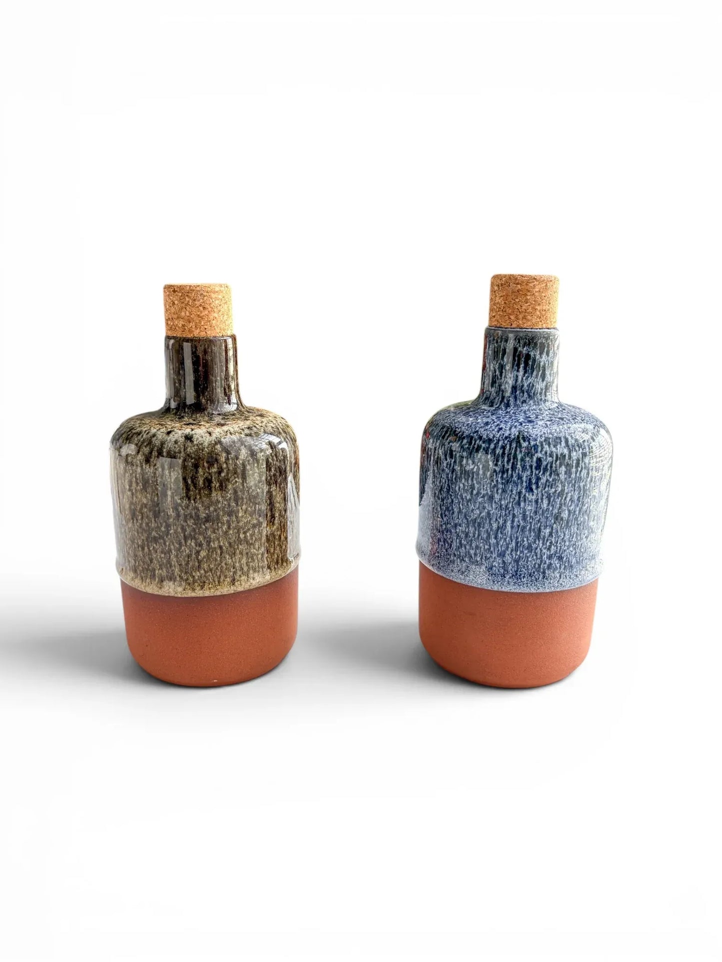 Two Wiid Design half-body ceramic olive oil bottles on a white background. One is finished in cobalt blue reactive glaze and the other in brown, both featuring raw terracotta bases and natural cork stoppers.