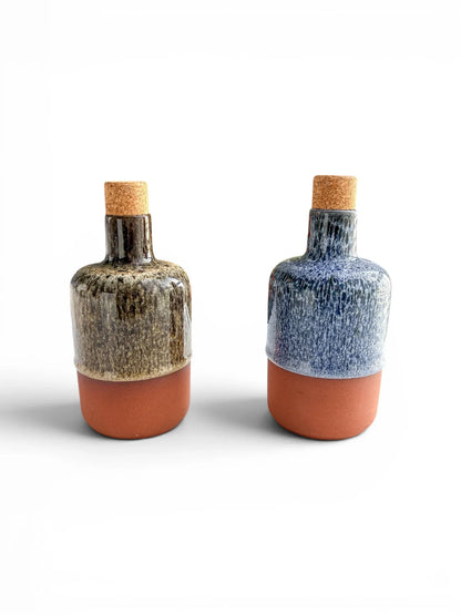 Two Wiid Design half-body ceramic olive oil bottles on a white background. One is finished in cobalt blue reactive glaze and the other in brown, both featuring raw terracotta bases and natural cork stoppers.
