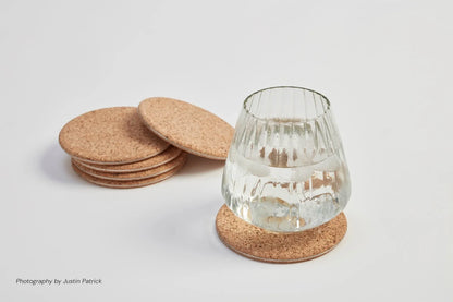 Lifestyle shot of the light natural cork and steel coasters. A ribbed glass of water with ice sits on one coaster, with a neat stack of matching light cork coasters in the background, highlighting the organic texture and 7mm thickness.