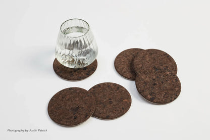 Lifestyle shot of the dark composite cork and steel coasters. A glass of water is placed on one dark, textured coaster, with four others fanned out on a white surface, displaying the rich, charred tones and the crisp industrial steel edge.