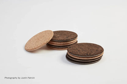 A comparison shot of Wiid Design natural cork and steel coasters. A single light composite cork coaster leans against two separate stacks of dark composite cork coasters, showcasing the visible stainless steel core and the tonal difference between the two natural materials.
