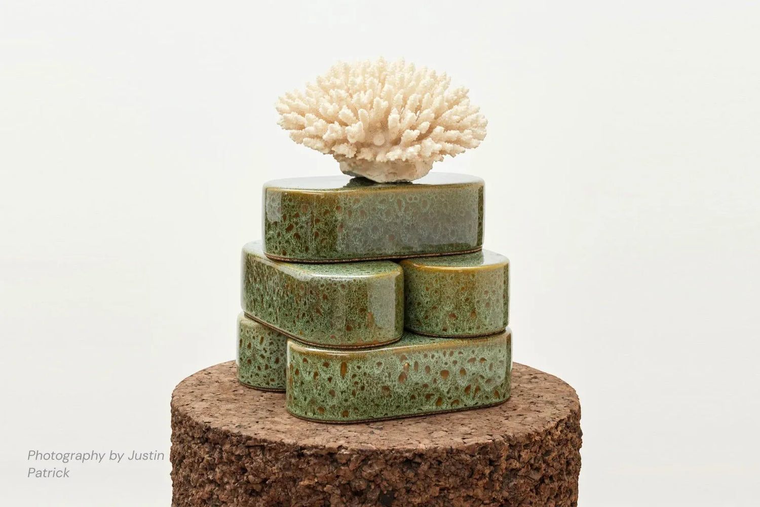 A creative lifestyle shot featuring a stack of five Wiid Design rounded ceramic bricks in reactive green glaze. The bricks are arranged on a large cylindrical dark cork base, topped with a piece of natural white coral to demonstrate their use as sculptural plinths.