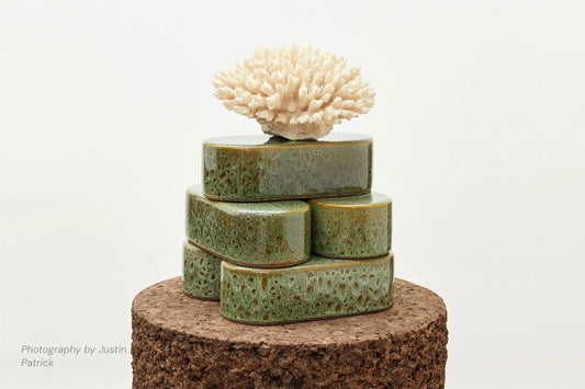 A creative lifestyle shot featuring a stack of five Wiid Design rounded ceramic bricks in reactive green glaze. The bricks are arranged on a large cylindrical dark cork base, topped with a piece of natural white coral to demonstrate their use as sculptural plinths.