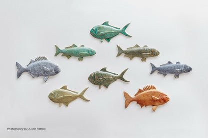 A curated wall display of eight Wiid Design sculptural ceramic fish on a white background. The collection features various species and reactive glazes, including shades of forest green, emerald, cobalt, and warm brown, arranged in a shoal-like formation that highlights the different textures and silhouettes.