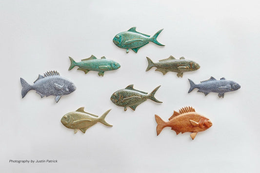 A curated wall display of eight Wiid Design sculptural ceramic fish on a white background. The collection features various species and reactive glazes, including shades of forest green, emerald, cobalt, and warm brown, arranged in a shoal-like formation that highlights the different textures and silhouettes.