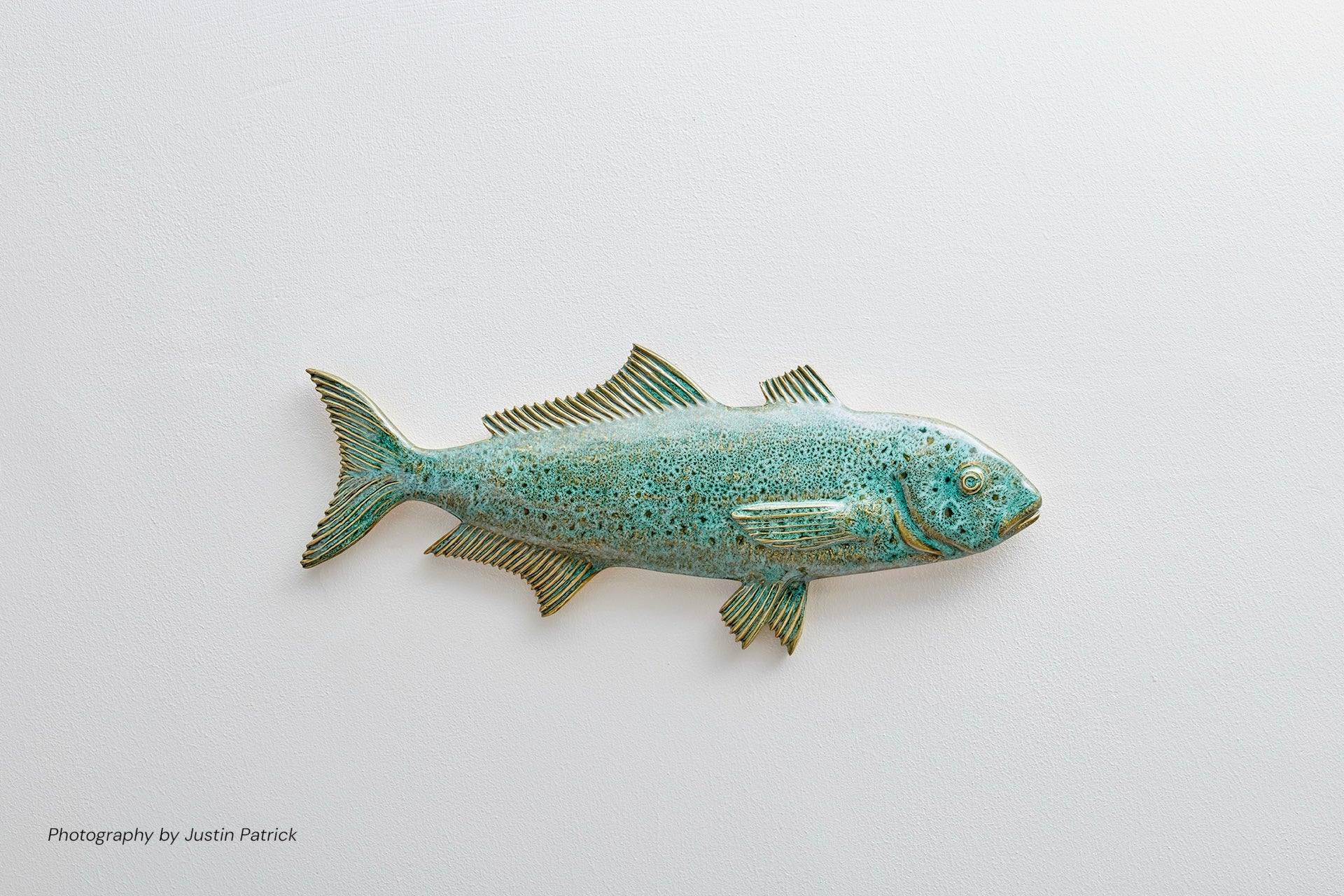 A side-profile view of the Wiid Design sculptural Ice Fish in emerald green reactive glaze. The high-gloss finish shows a fine, dark speckle and variegated tones over the detailed scales and fins, mounted against a light, neutral wall.