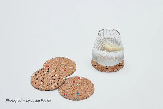 Lifestyle shot of Wiid Design speckled cork and steel coasters on a white background. One coaster holds a ribbed glass of water with a lemon slice, while a set of three coasters in the multi-colour and black-and-white variants are scattered nearby.