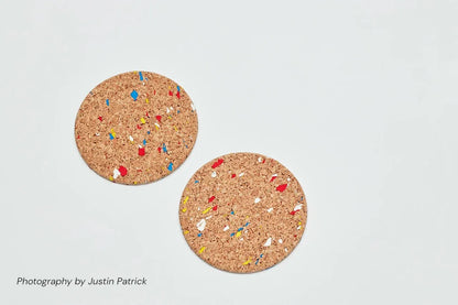 Top-down view of two Wiid Design cork coasters in the multi-colour speckled variant. The natural light cork is scattered with recycled rubber flecks in vibrant blue, red, and yellow, creating a terrazzo-style finish.