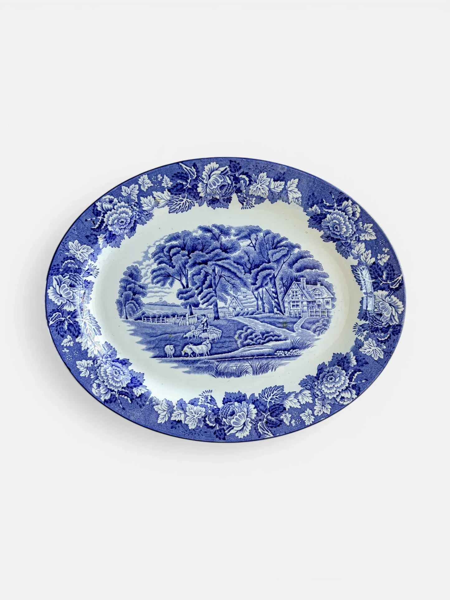 A top-down, professional product photograph of an oval Wood & Sons "English Scenery" blue and white transferware platter. The platter features a central pastoral scene of a cottage, sheep, and large trees, framed by a wide, dense border of peonies and foliage against a crisp white background.