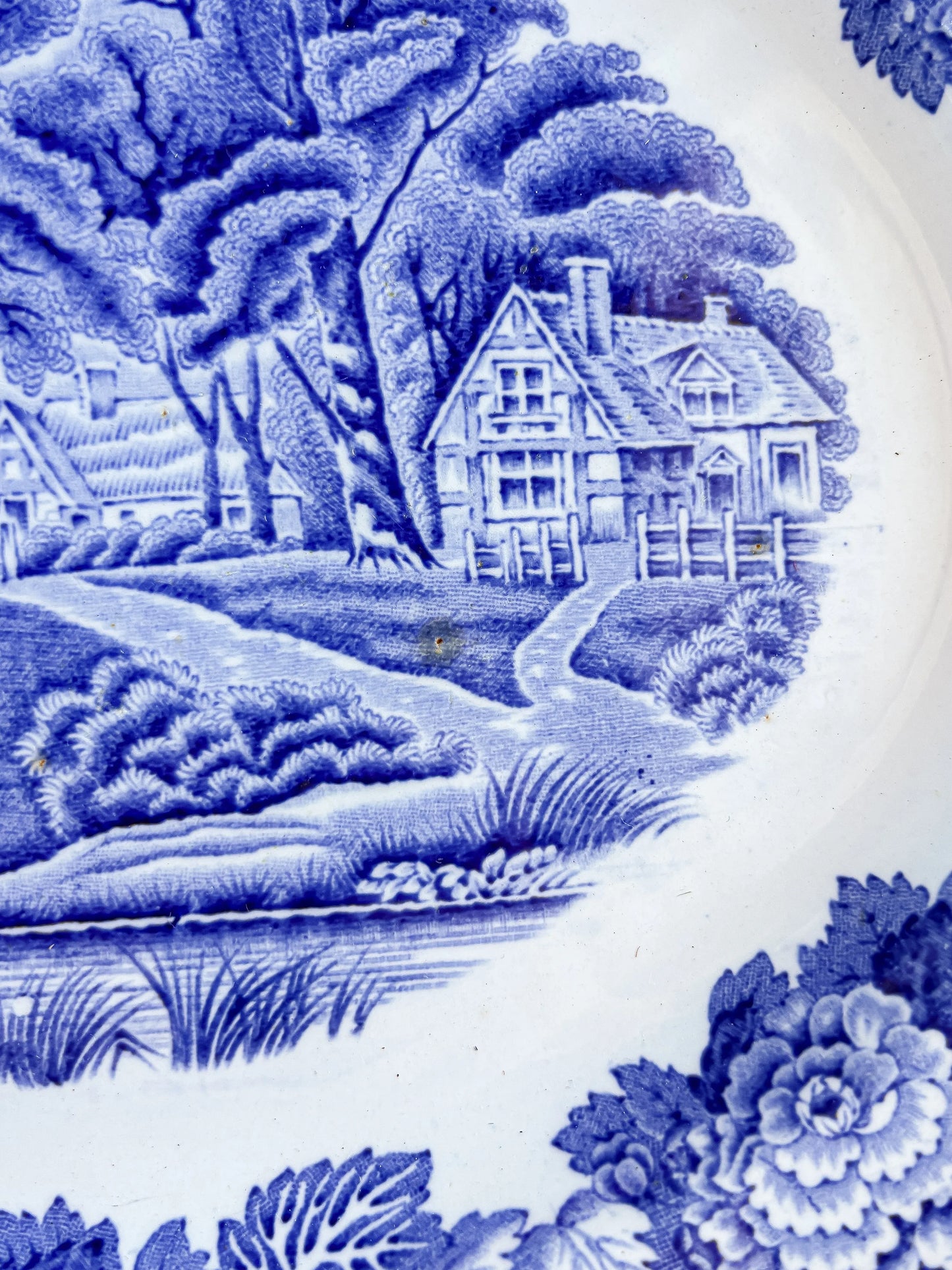 A detailed close-up of the "English Scenery" transferware pattern, highlighting the fine blue lines of a Tudor-style cottage, rustic fencing, and lush shrubbery. A small, dark brown iron spot is visible in the centre of the scene, characteristic of antique earthenware.