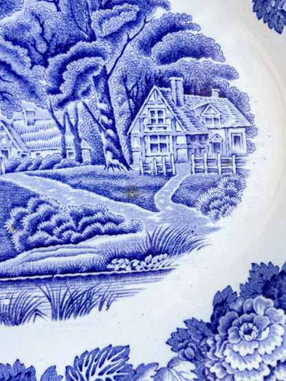 A detailed close-up of the "English Scenery" transferware pattern, highlighting the fine blue lines of a Tudor-style cottage, rustic fencing, and lush shrubbery. A small, dark brown iron spot is visible in the centre of the scene, characteristic of antique earthenware.